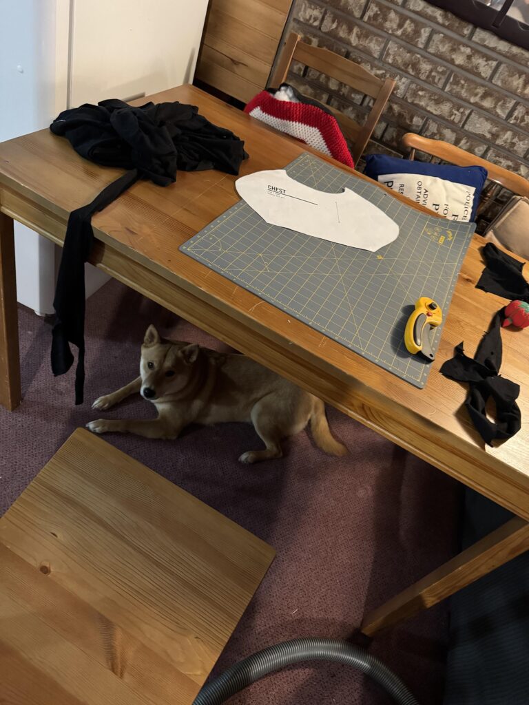 A Shiba Inu under a table with a cutting mat and the pattern and a pile of spandex